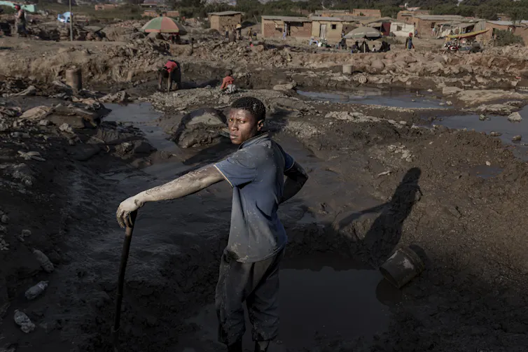 A man with dried mud on his bare arms stand near a water-filled mine where a child and woman are searching for minerals.